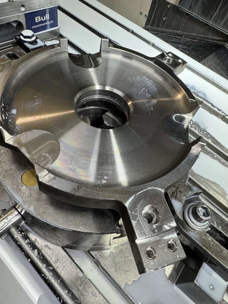 Nahaufnahme einer Metallbearbeitungsmaschine mit einer runden Drehscheibe und Werkzeugen. Die Maschine ist mit Kühlmittel bedeckt, was auf einen aktiven Bearbeitungsprozess hindeutet.