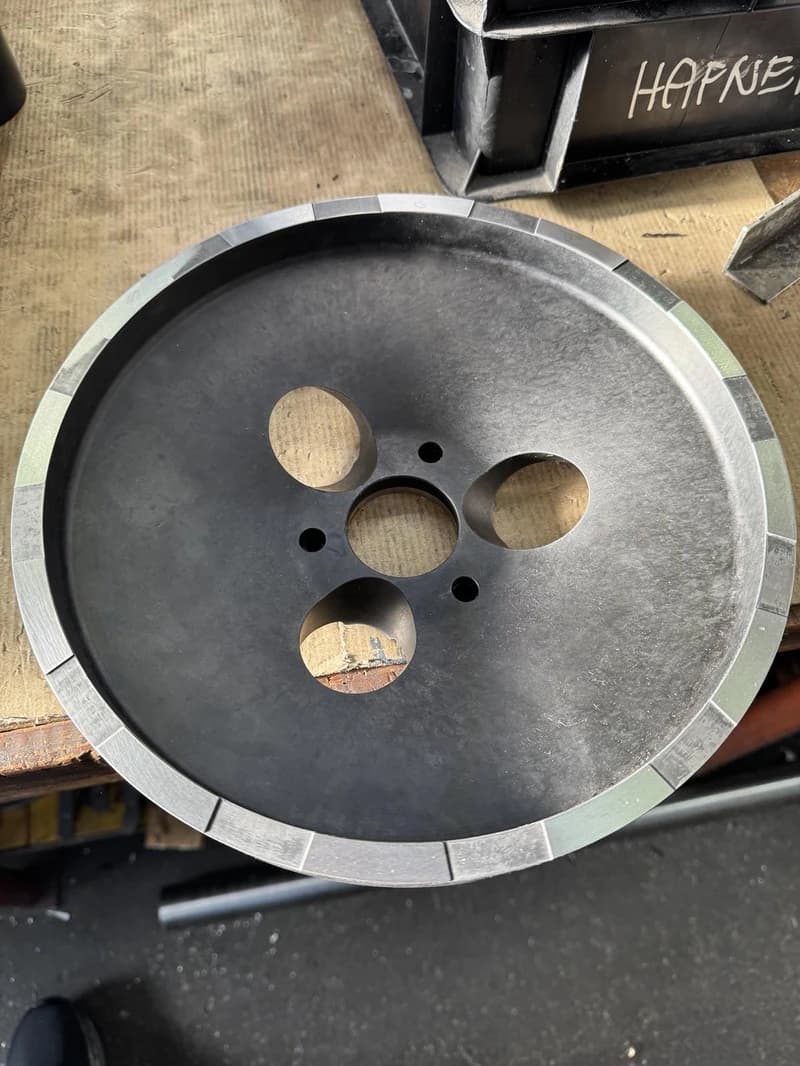 Ein graues Metallrad mit mehreren Loechern liegt auf einer Werkbank. Das Rad hat einen segmentierten Rand.