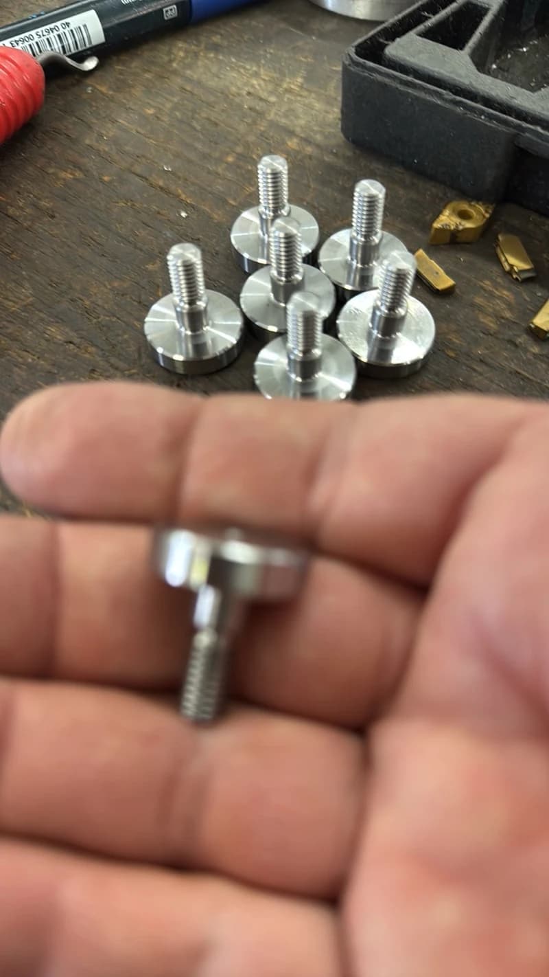 Nahaufnahme von mehreren silbernen Metallschrauben mit flachem Kopf, von denen eine in einer Hand gehalten wird. Die Schrauben liegen auf einer Holzoberfläche, zusammen mit einem schwarzen Behälter und einigen goldenen Werkzeugspitzen.