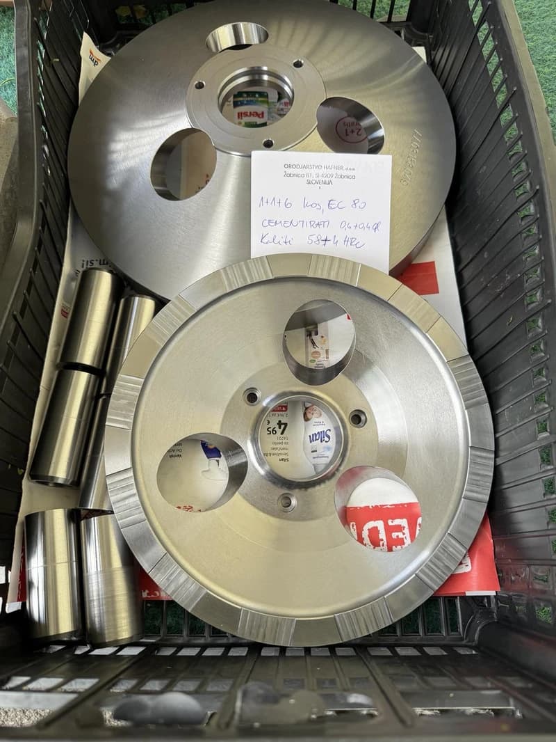 Zwei metallische Scheiben mit Löchern und zylindrische Metallteile liegen in einem schwarzen Korb. Ein Zettel mit handschriftlichen Notizen liegt auf einer der Scheiben.